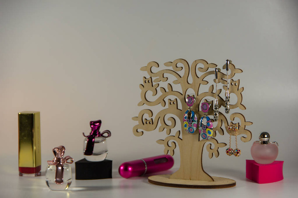 Holzständer mit drei Paar langen Ohrringen, geschlossenem Lippenstift in Goldfarbe, schwarzen und rosa Schwämmen, kleinen Fläschchen mit duftendem Parfüm auf weißem Hintergrund. - Foto, Bild