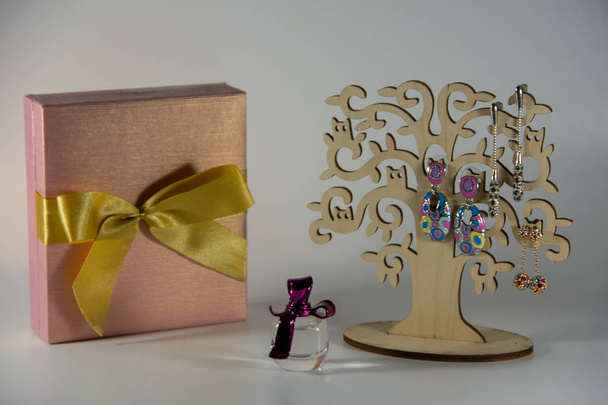 A beautiful stand in the form of a small tree with long earrings, a small bottle of fragrant perfumes, a pink gift box decorated with a gold bow and on a white background. - Photo, Image