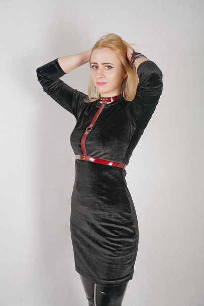 Beautiful young blonde woman in velvet black dress and red leather harness over clothes posing in white studio - Zdjęcie, obraz