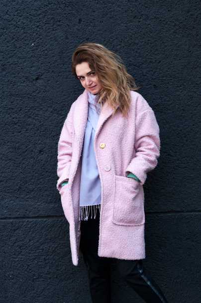 beautiful smiling woman with long hair in pink coat, purple scarf and pants looking at camera and holding hands in pockets on black background - Foto, immagini