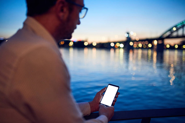 Бизнесмен, использующий мобильный телефон возле реки с городским освещением в б
 - Фото, изображение
