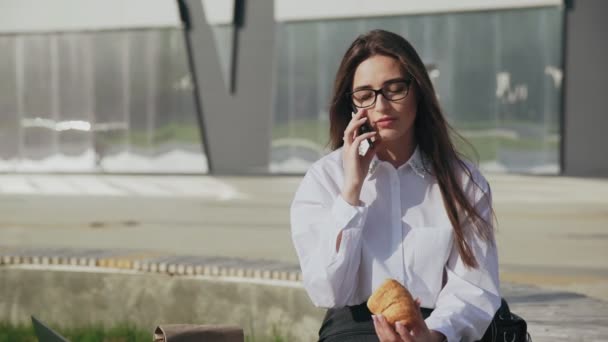 Kobieta sukcesu korporacji jedząca rogalika podczas rozmowy przez telefon komórkowy i uśmiechająca się na świeżym powietrzu - Materiał filmowy, wideo