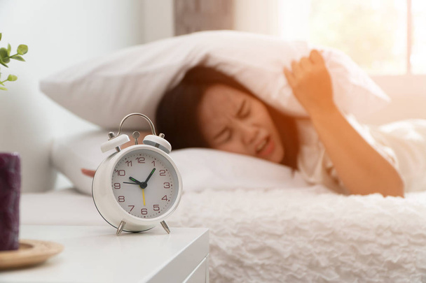 Ospalá asijská žena spí na posteli a používá polštář k zavření ucha, aby neslyšel Budík v ložnici. Koncept Lenošného dne a chci spát a nechci se ráno probudit - Fotografie, Obrázek