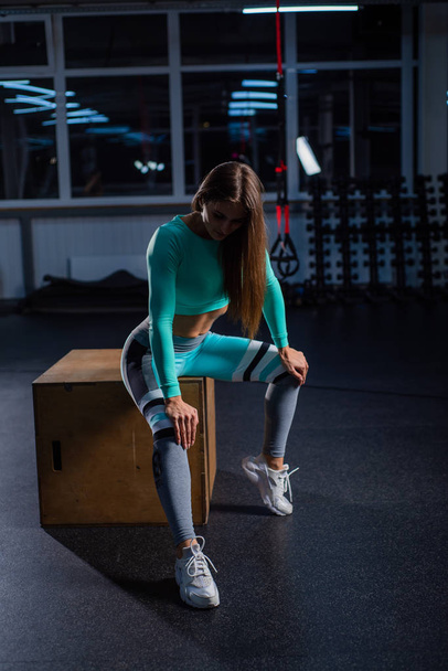 A beautiful woman in a dark room sits on a cube. The coach is resting after a productive roundabout. Female athlete in sportswear in the gym. - Fotografie, Obrázek