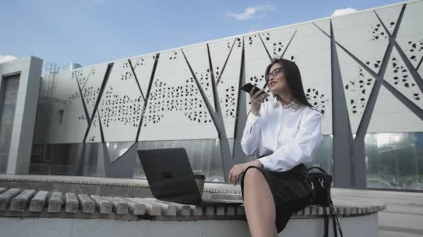 Obchodní žena nahrává hlasovou zprávu, zatímco sedí v parku se svým notebookem - Záběry, video