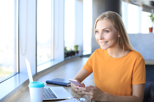Kaunis liiketoiminnan nainen käyttää älypuhelinta hymyillen istuessaan työpaikallaan
 - Valokuva, kuva