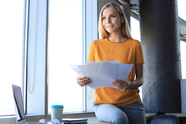 Attractive business woman holding documents and looking at them while sitting on the desk in office - Foto, Imagen