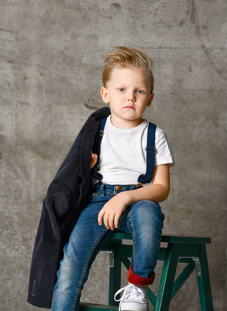 Cute stylish boy sitting on chair near concrete wall - Фото, изображение