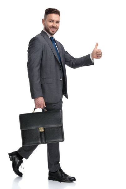 χαμογελαστός νεαρός επιχειρηματίας περπατάει με μια βαλίτσα και κάνει εντάξει - Φωτογραφία, εικόνα