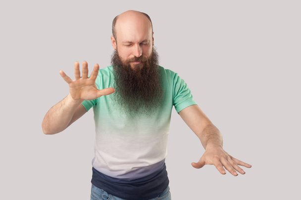 f middle aged bald man with beard in light green t-shirt standing with closed eyes and trying to touching on grey background. - Fotoğraf, Görsel