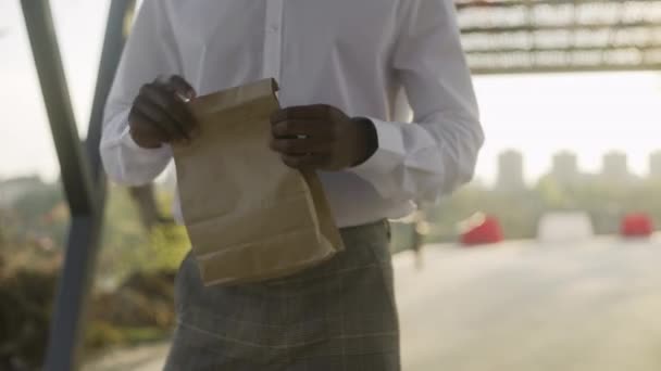 Młody afrykański biznesmen w białej koszuli wziął rogalika z paczki - Materiał filmowy, wideo