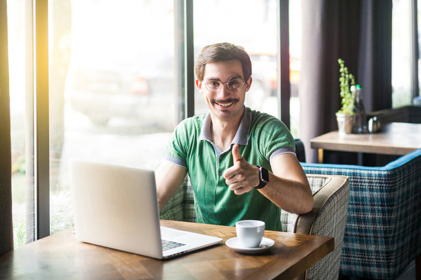 Young happy businessman in green t-shirt working on laptop and showing thumb up at camera while sitting at table in cafe, freelancing concept - Фото, зображення