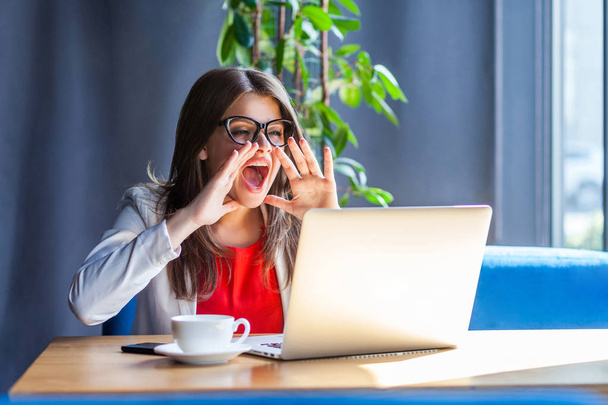 angry brunette young woman in glasses looking at laptop screen on video call and screaming while sitting at table in cafe, freelancing concept  - Valokuva, kuva