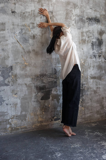 lonely brunette girl in white blouse and black jeans stands on tiptoe, stretches and leans against wall in emty room - Photo, Image