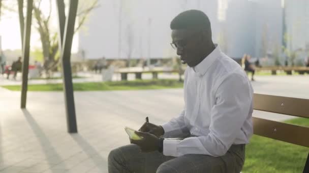 Junger Afroamerikaner auf einer Parkbank schreibt seine Gedanken in ein Tagebuch - Filmmaterial, Video