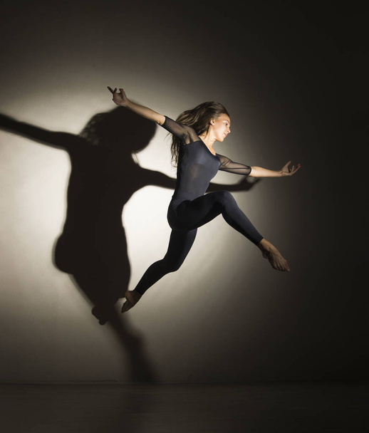 Girl in dark performs gymnastic jumping, on a white background there is a shadow from a shape. - Valokuva, kuva