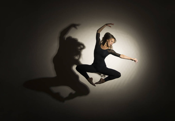 Girl in dark performs gymnastic jumping, on a white background there is a shadow from a shape. - Fotoğraf, Görsel