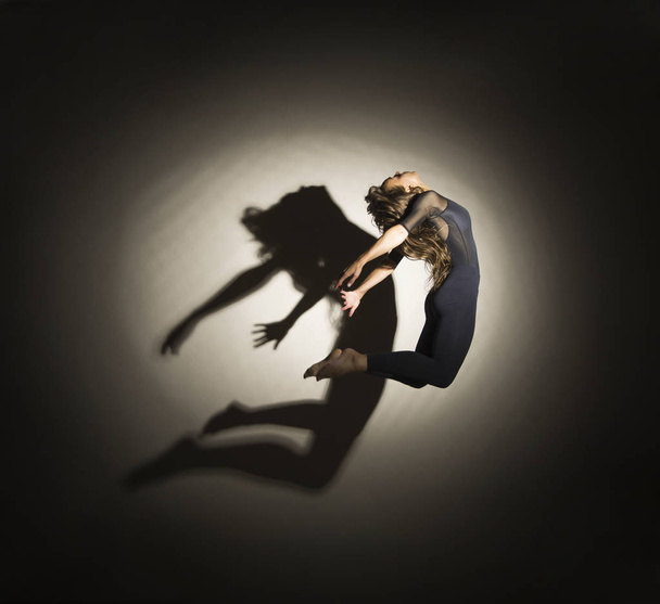 Girl in dark performs gymnastic jumping, on a white background there is a shadow from a shape. - Photo, Image