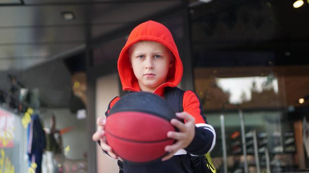 Nuori poika harjoituspuvussa poseeraa koripallon kanssa kaupan edessä
. - Valokuva, kuva