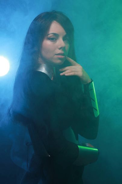 φουτουριστικό μοντέλο φορώντας μαύρα και ασημένια ρούχα και στέκεται στο πολύχρωμο μπλε και πράσινο καπνό - Φωτογραφία, εικόνα