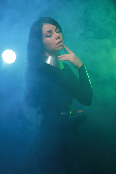 futuristic fashion model wearing black and silver clothes and standing in the colorful blue and green smoke - Photo, image