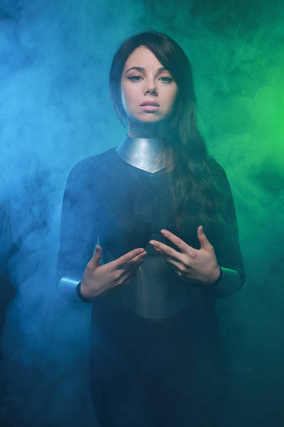 futuristinen malli yllään musta ja hopea vaatteita ja seisoo värikäs sininen ja vihreä savu
 - Valokuva, kuva
