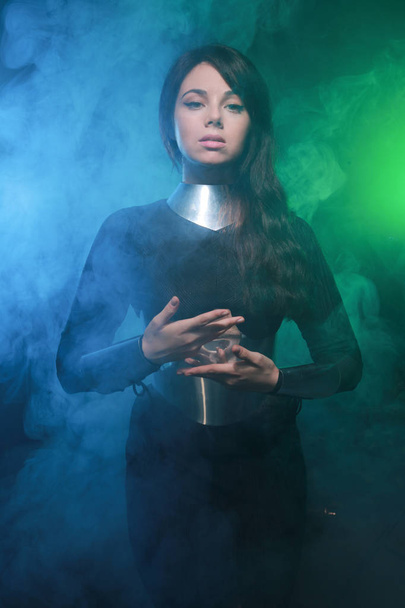 futuristisches Mode-Model trägt schwarze und silberne Kleidung und steht im farbenfrohen blauen und grünen Rauch - Foto, Bild