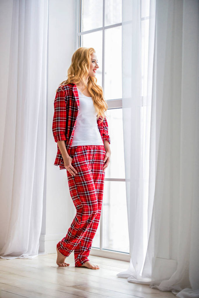 赤いチェッカーのパジャマの美しいブロンドの女性は、窓の近くに立って、快適な家の服を示しています - 写真・画像