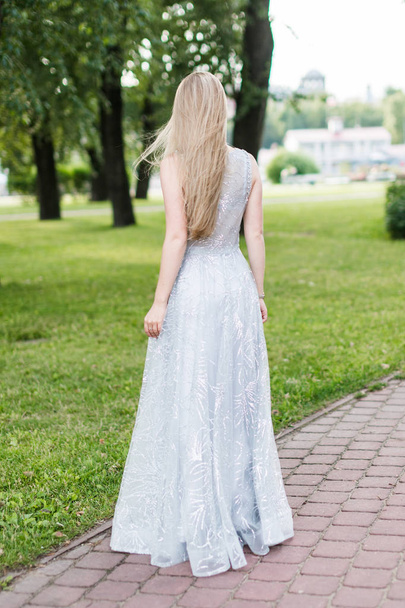 Slender blonde girl in grey beaded floor-length dress turned her back in park. - Zdjęcie, obraz
