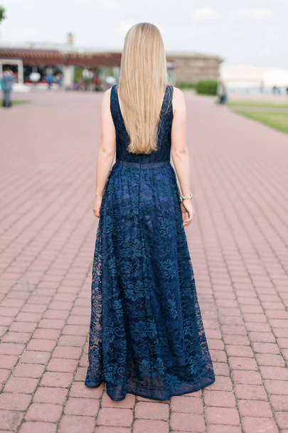 Slender blonde girl in dark-blue floor-length dress turned her back on the pavement. - Photo, Image