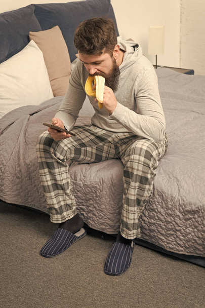 brutal sleepy man has breakfast in bedroom. mature male with beard in pajama on bed. sleep and awake. healthy lifestyle. bearded man hipster eat banana in morning. Chatting with girlfriend - 写真・画像