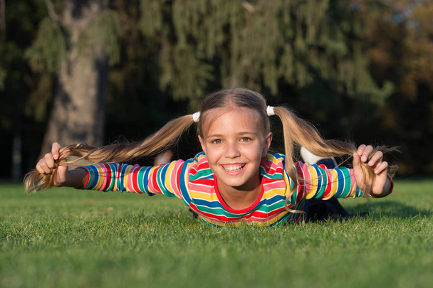 Cheerful schoolgirl on sunny day. Happy smiling pupil. Have fun. Girl kid laying green grass. Healthy emotional happy kid relaxing outdoors. Girl ponytails hairstyle enjoy relax. Living happy life - Foto, imagen