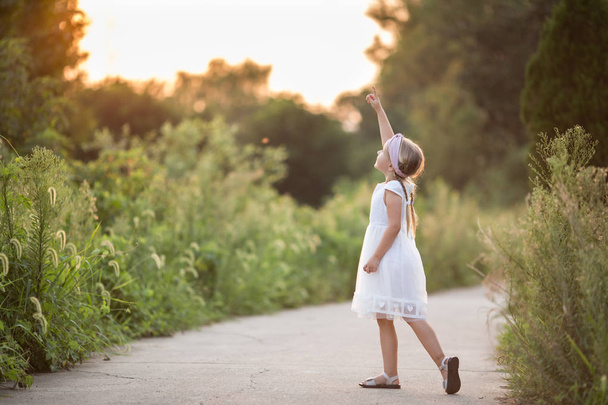 Портрет очаровательной маленькой девочки с длинными светлыми волосами в парке солнечный летний день. Парень показывает что-то с пальцем на улице. Прекрасное дитя. Счастливый улыбчивый малыш, день детства, концепция образования
 - Фото, изображение