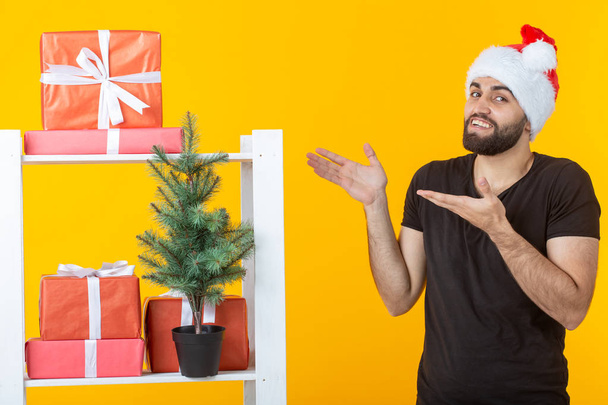 Sakallı ve Noel Baba şapkalı genç pozitif adam sarı arka planda hediyeler ve Noel ağacı ile standı yakınında duruyor. Noel ve Yeni Yıl için tebrik hediyesi ve indirimler kavramı. - Fotoğraf, Görsel