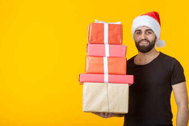 Disgruntled young man with a beard in a Santa Claus hat holds five gift boxes posing on a yellow background with copyspace. Concept of gifts and greetings for Christmas and New Year. - Foto, imagen