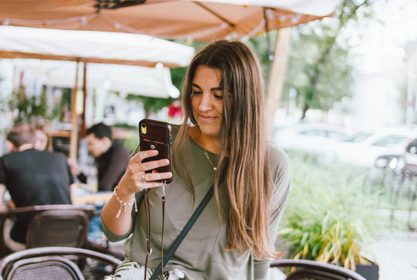 Joven sonriente pelo largo morena chica vestida casual usando móvil en la calle cafetería
 - Foto, imagen