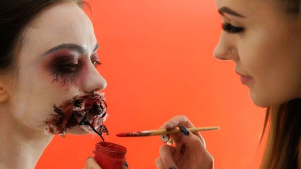 Bodypainting. der Prozess, gruselige Schminke aufzutragen, um Halloween zu feiern. Das Mädchen klebt blutige Farbe auf den genähten Mund eines anderen Mädchens. roter Hintergrund. - Foto, Bild