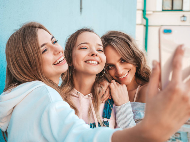 Three young smiling hipster women in summer clothes.Girls taking selfie self portrait photos on smartphone.Models posing in the street near wall.Female showing positive face emotions - Photo, image