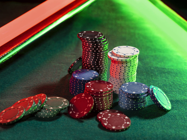 Close-up foto van een veelkleurige chips palen, sommige van hen liggen in de buurt op de groene cover van de speeltafel, onder groene en rode neon lichten. Zwarte achtergrond. Close-up. - Foto, afbeelding