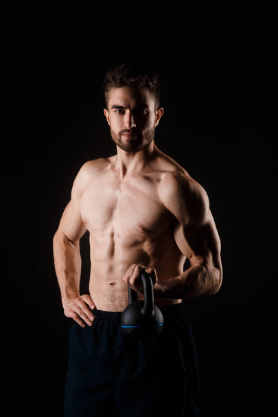 Young bearded man with a kettlebell in his hands on a black background. bare-chested athlete - Foto, Bild