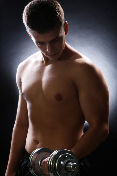 Handsome young muscular sportsman execute exercise with dumbbell on dark background - Fotografie, Obrázek