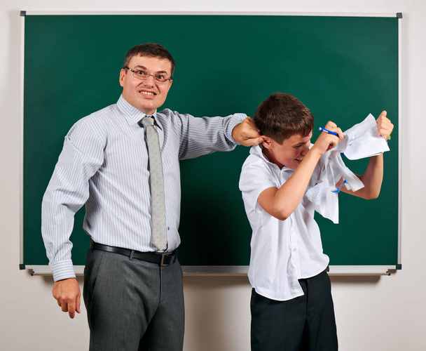 Das Porträt eines Lehrers erregt das Ohr lustiger Schüler mit geringer Disziplin. Schüler sehr emotional, lustig und sehr glücklich, posiert vor Tafel-Hintergrund - zurück zum Schul- und Bildungskonzept - Foto, Bild