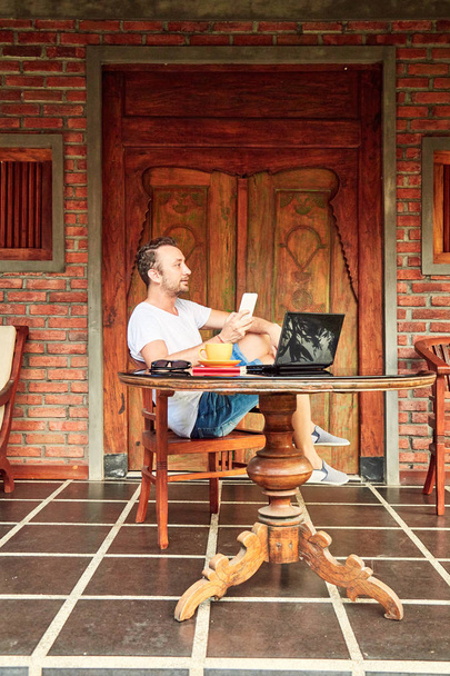 Man met smartphone, koffie/thee en laptop op een thuis veranda. - Foto, afbeelding
