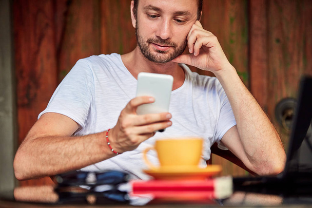Hombre con teléfono inteligente, café / té y portátil en un porche de casa
. - Foto, Imagen