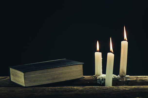 Hermosa biblia santa cerrada y tres velas blancas quemadas sobre una vieja mesa de madera de roble y fondo negro
.  - Foto, Imagen