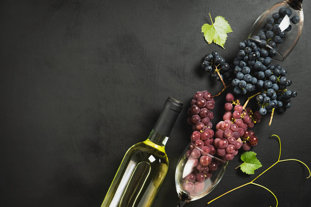 White wine bottle, wineglasses, fresh grapes and leaves on black wood background. Flat lay, top view, copy space. Wine bar, winery, wine tasting concept - Φωτογραφία, εικόνα