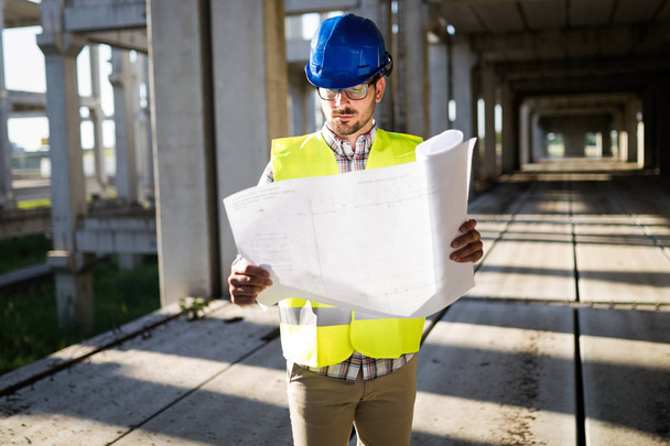 Portrait of male site contractor engineer with hard hat holding blue print paper at construction site, - Zdjęcie, obraz