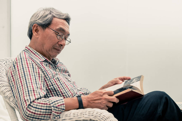 Senior Asian man reading book on chair at home. - Photo, Image