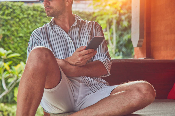 Człowiek za pomocą inteligentnego telefonu na kanapie taras w lecie. - Zdjęcie, obraz