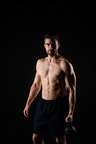 Young bearded man with a kettlebell in his hands on a black background. bare-chested athlete - Фото, изображение